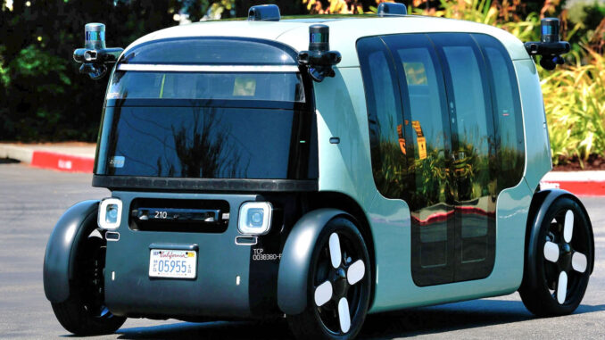 zoox in Las Vegas A futuristic, boxy, self-driving electric vehicle with large windows and black-and-white wheels is parked on a road with greenery in the background.
